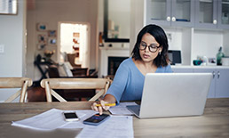 Woman sitting at kitchen table on her laptop and touching her cell phone.